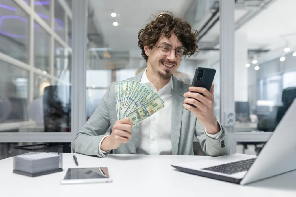 Successful young businessman with curly hair holding cash and using smartphone at office desk